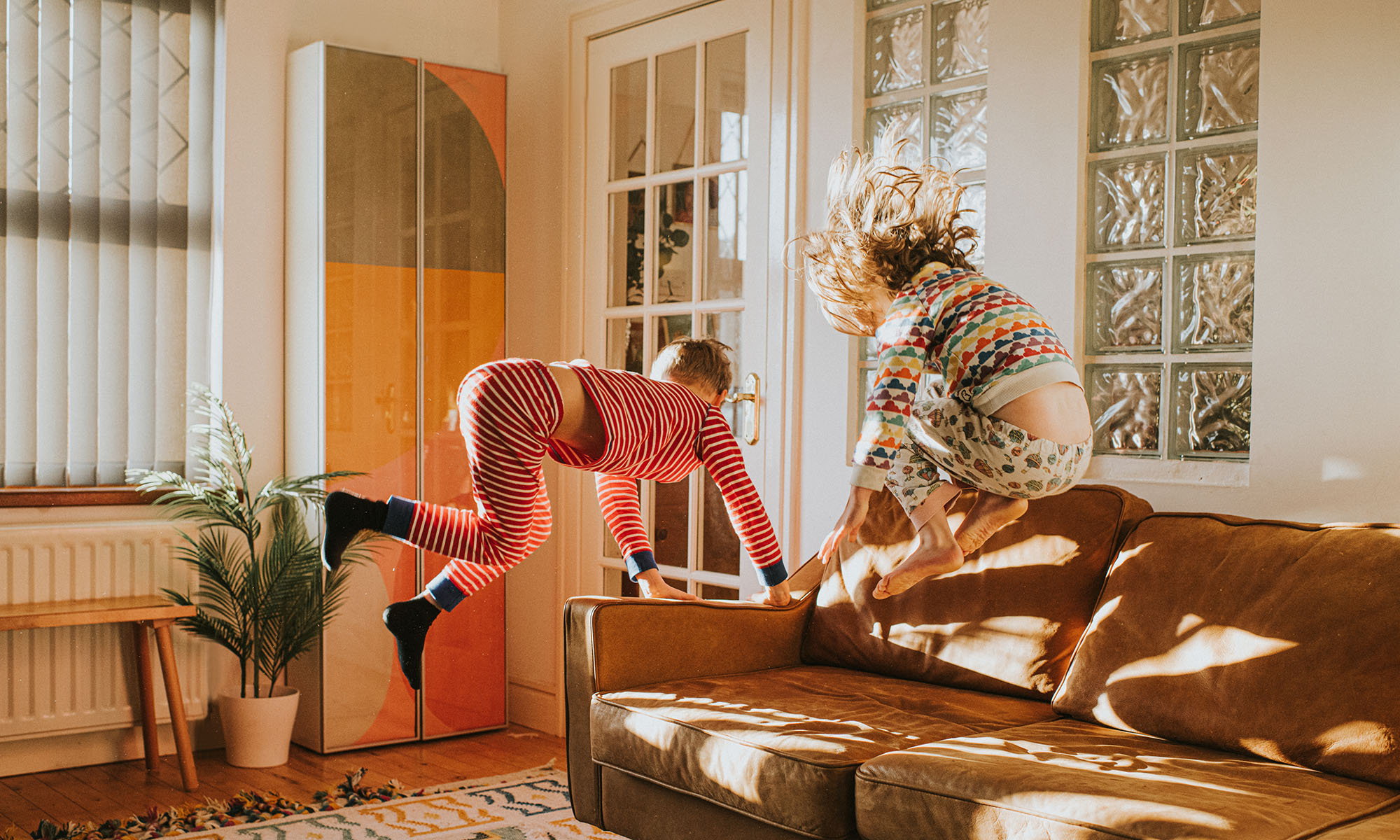 Two cute kids wearing pyjamas bounce on a brown leather couch in a sunny domestic room